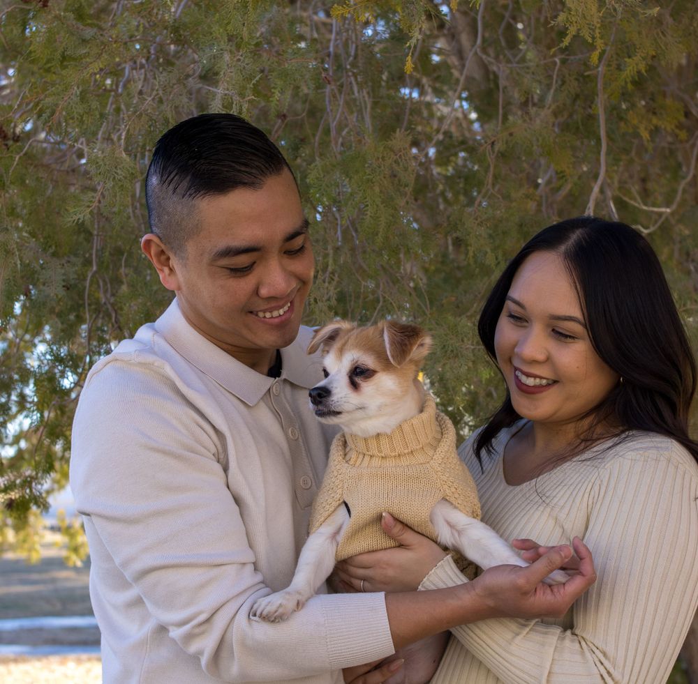 Happy couple smiling and holding their small dog in a yellow sweater under green trees outdoors.