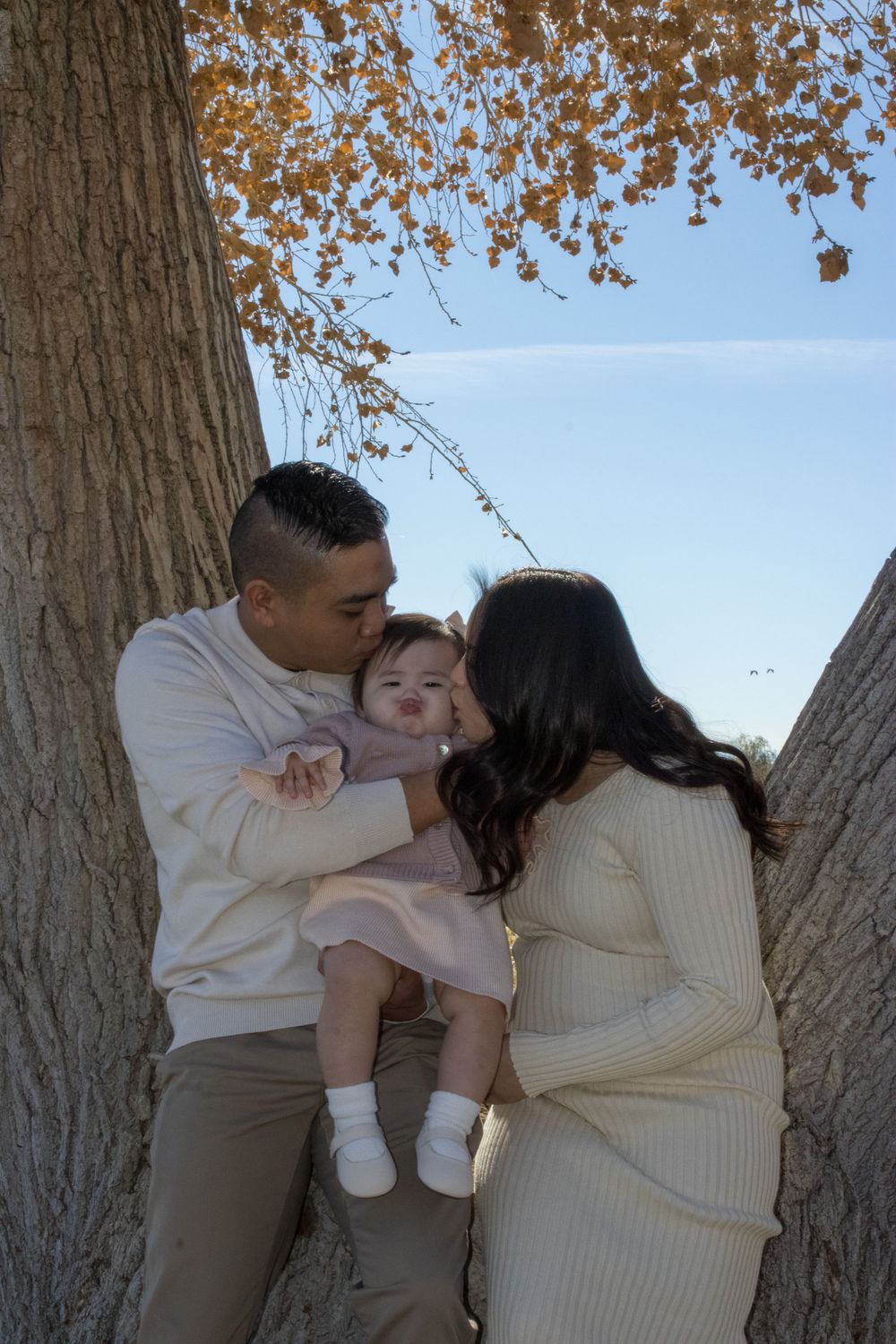 Loving parents in cream outfits kissing their baby girl between two large trees with fall foliage.