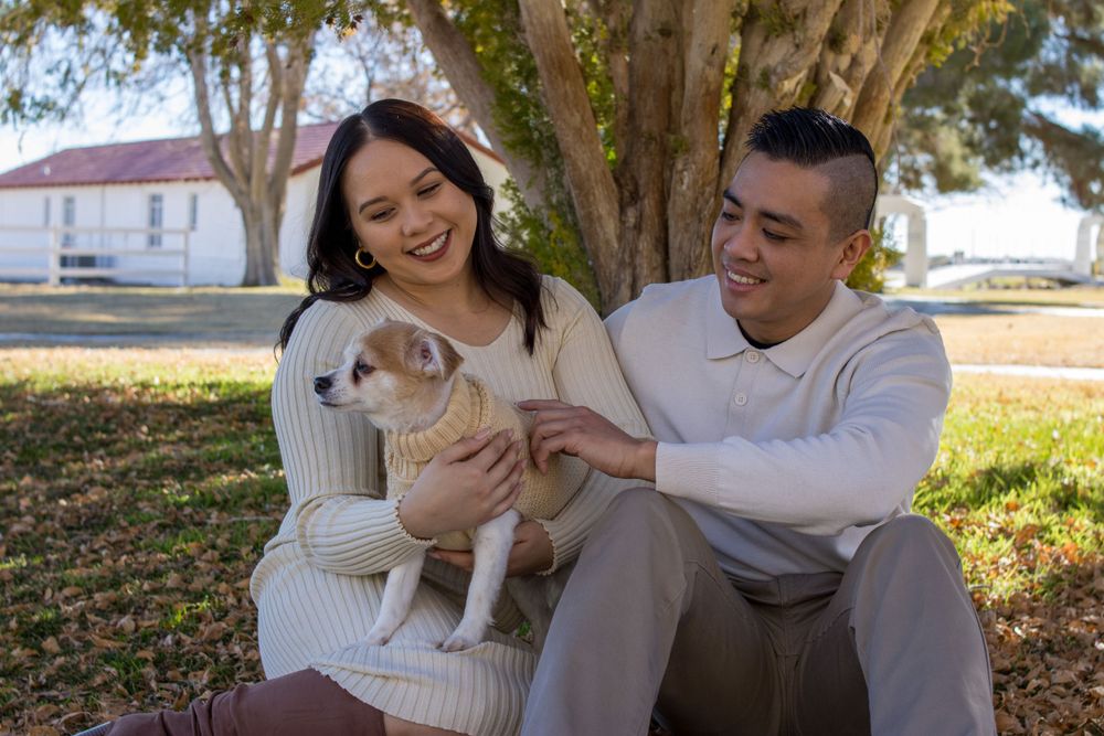 Happy couple sitting on grass with adorable puppy under a tree during fall season outdoors.