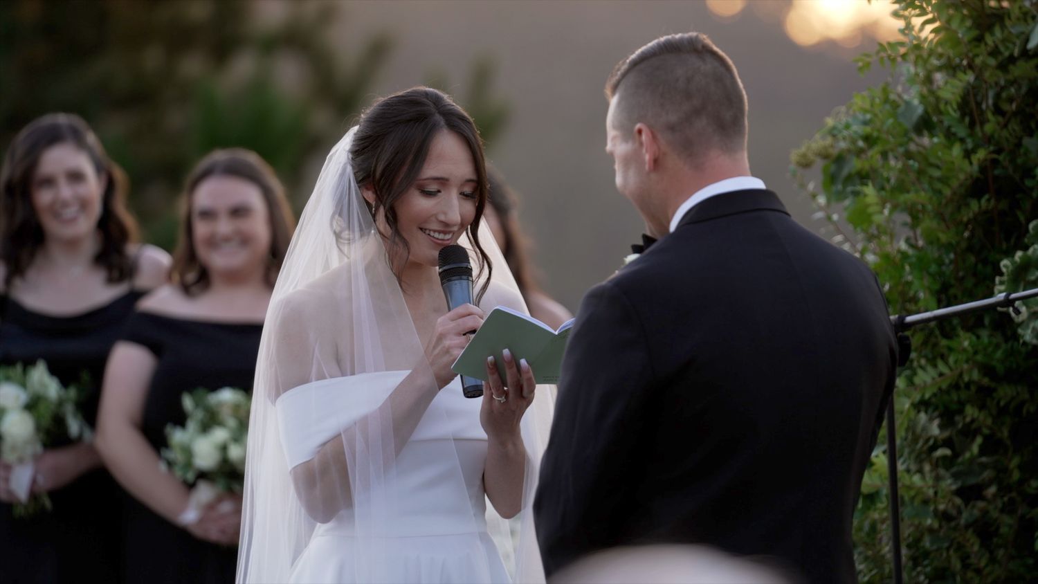 A bride reads her vows during an outdoor wedding ceremony at sunset while bridesmaids watch in the background.