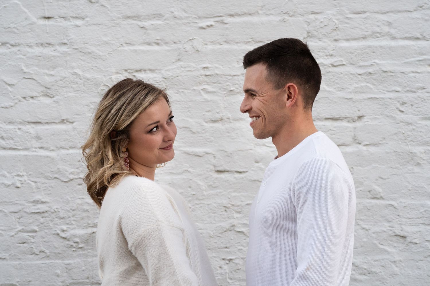 A couple in matching white sweaters poses against a white brick wall.