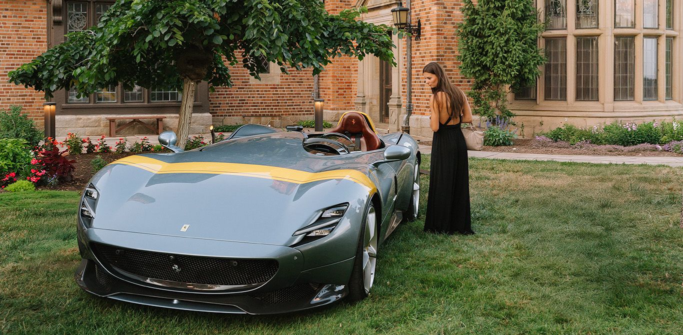 A woman in a black dress observing a Ferrari parked on the grass in front of Meadow Brook Hall