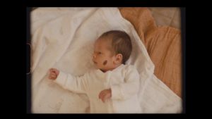 Newborn baby in white clothing stretches while resting on white bedding.