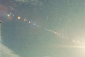 Sun flare creates a dreamy effect against a pale blue sky with scattered clouds, captured on super 8 film.
