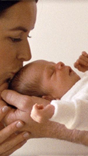 A tender moment as someone holds and gazes lovingly at a sleeping newborn baby wrapped in white.