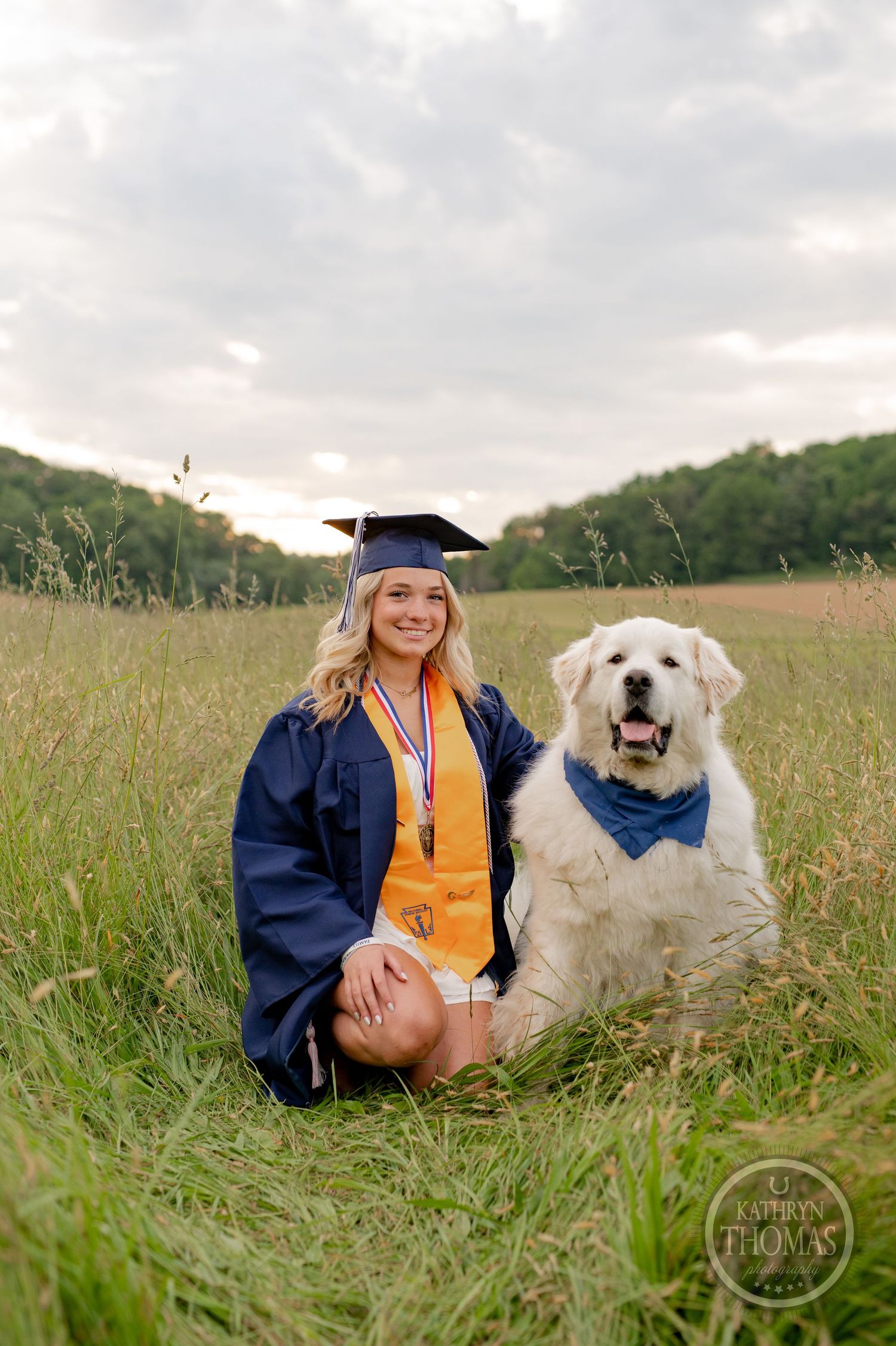 Graduate and fluffy white dog pose together in tall grass wearing matching navy graduation attire.