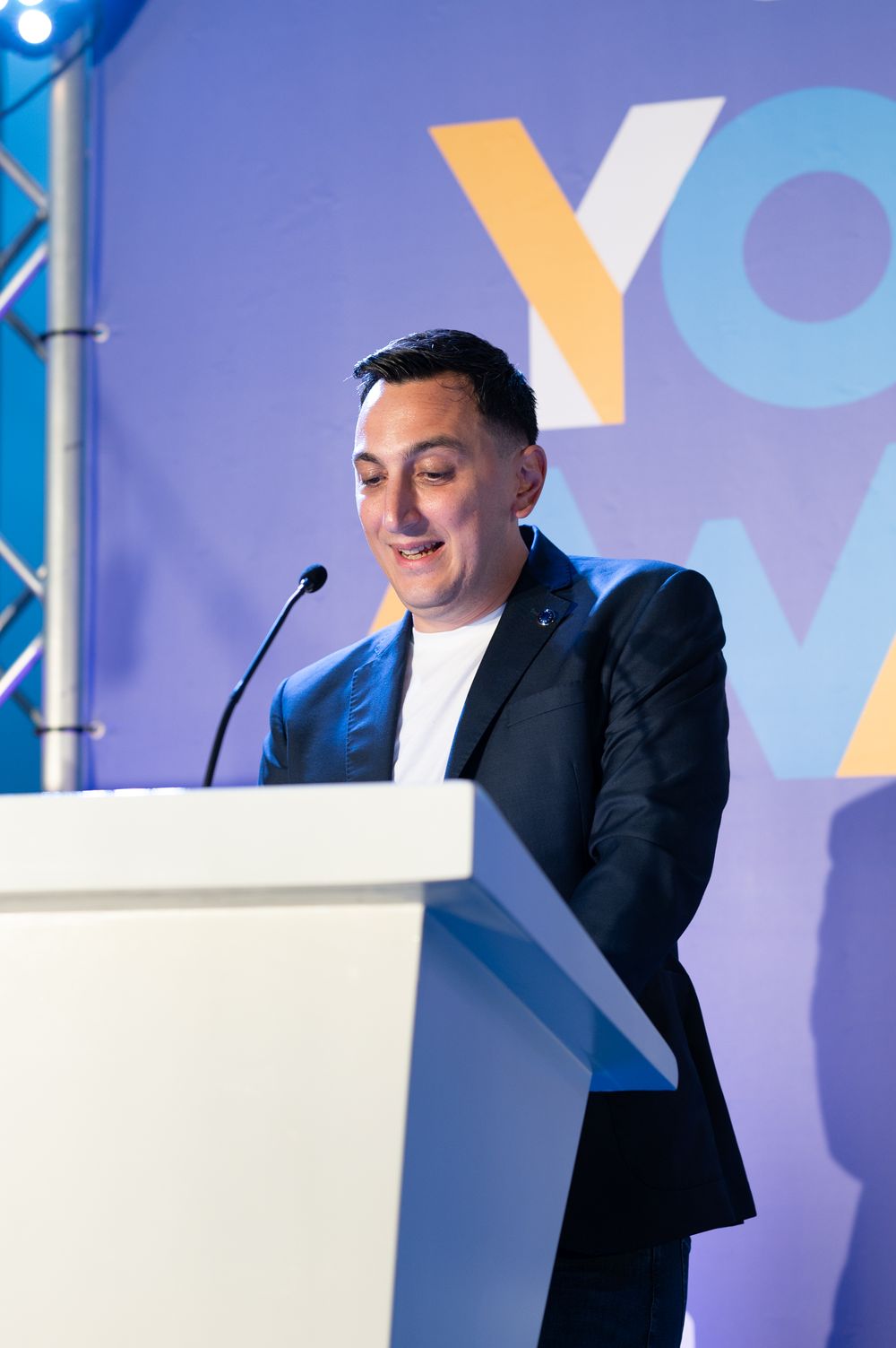 Professional speaker at white podium against blue geometric backdrop during presentation.