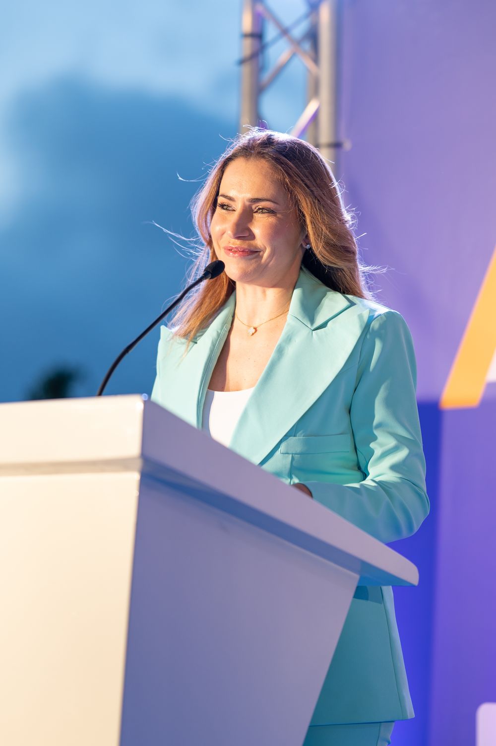 Presenter in turquoise suit jacket speaking at white podium during awards ceremony.