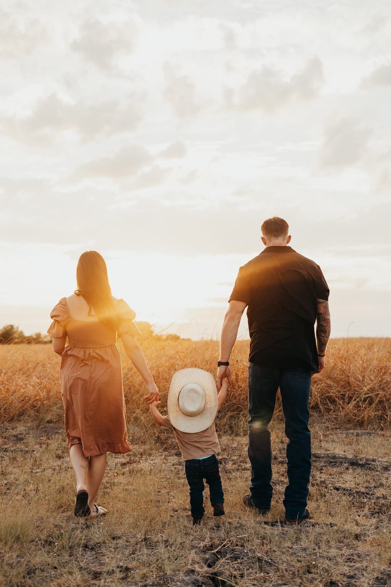 Family of three holding hands walking through golden wheat field toward dramatic sunset sky.