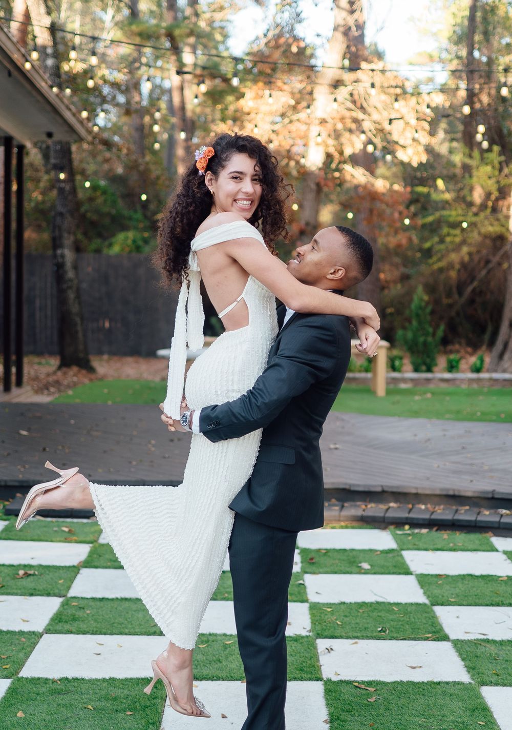 A couple in formal attire dances playfully on a giant outdoor checkerboard pattern in a park setting.