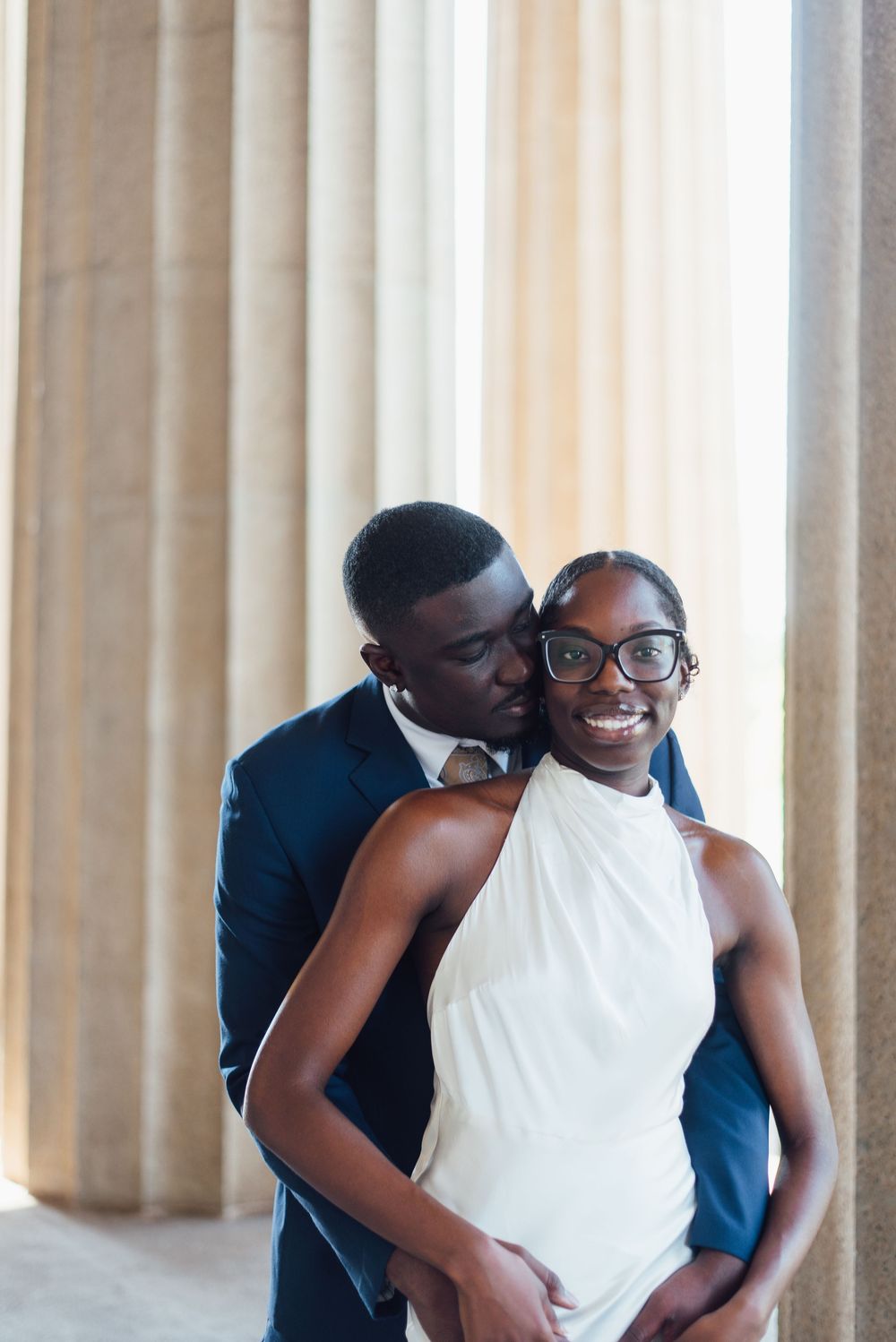 A romantic couple shares tender moments in front of elegant white columns while wearing formal attire.