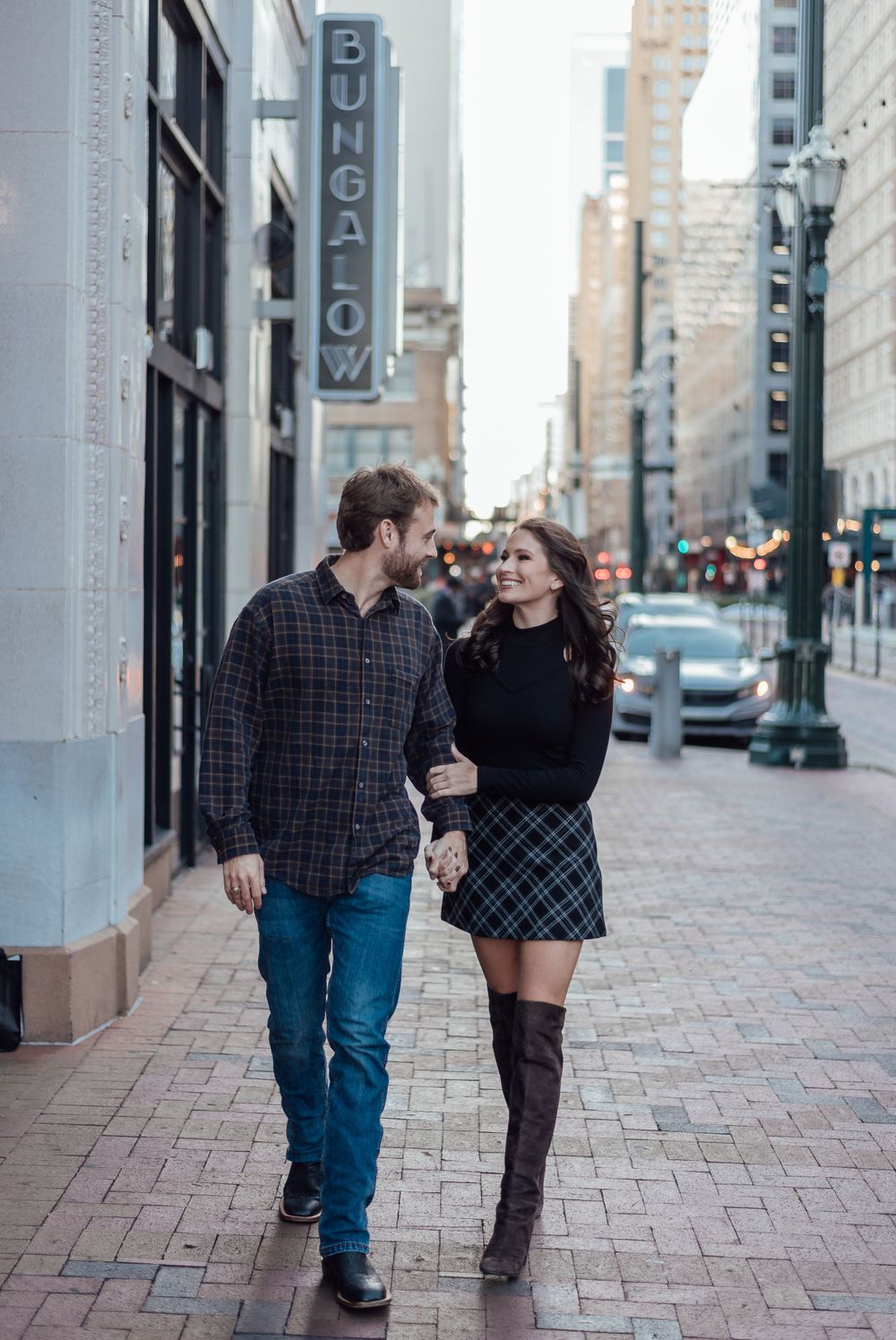 A couple walks together on a brick sidewalk in an urban setting with tall buildings and a vintage sign in the background.