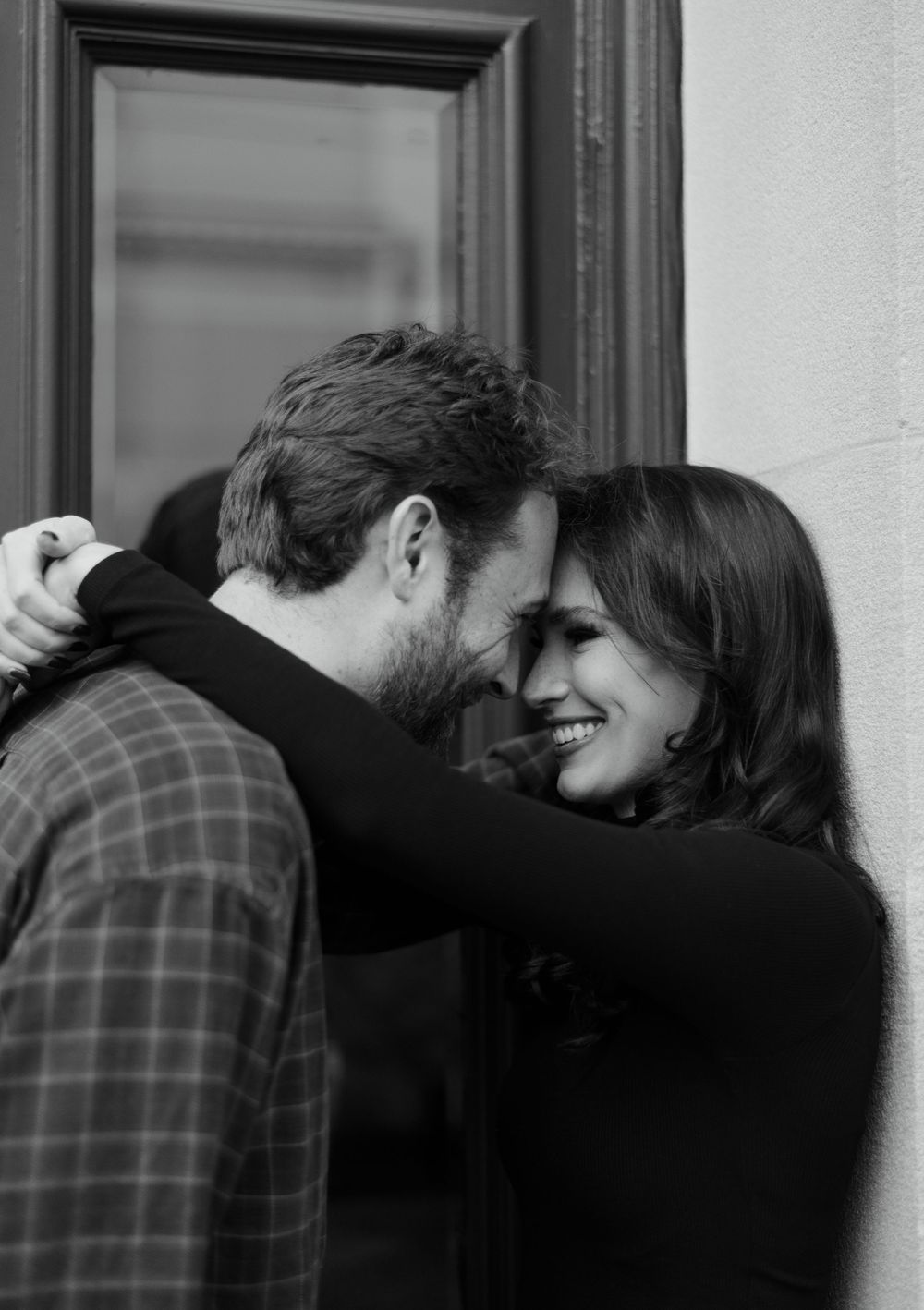 A romantic black and white photograph of a couple sharing an intimate moment while laughing against a wall.