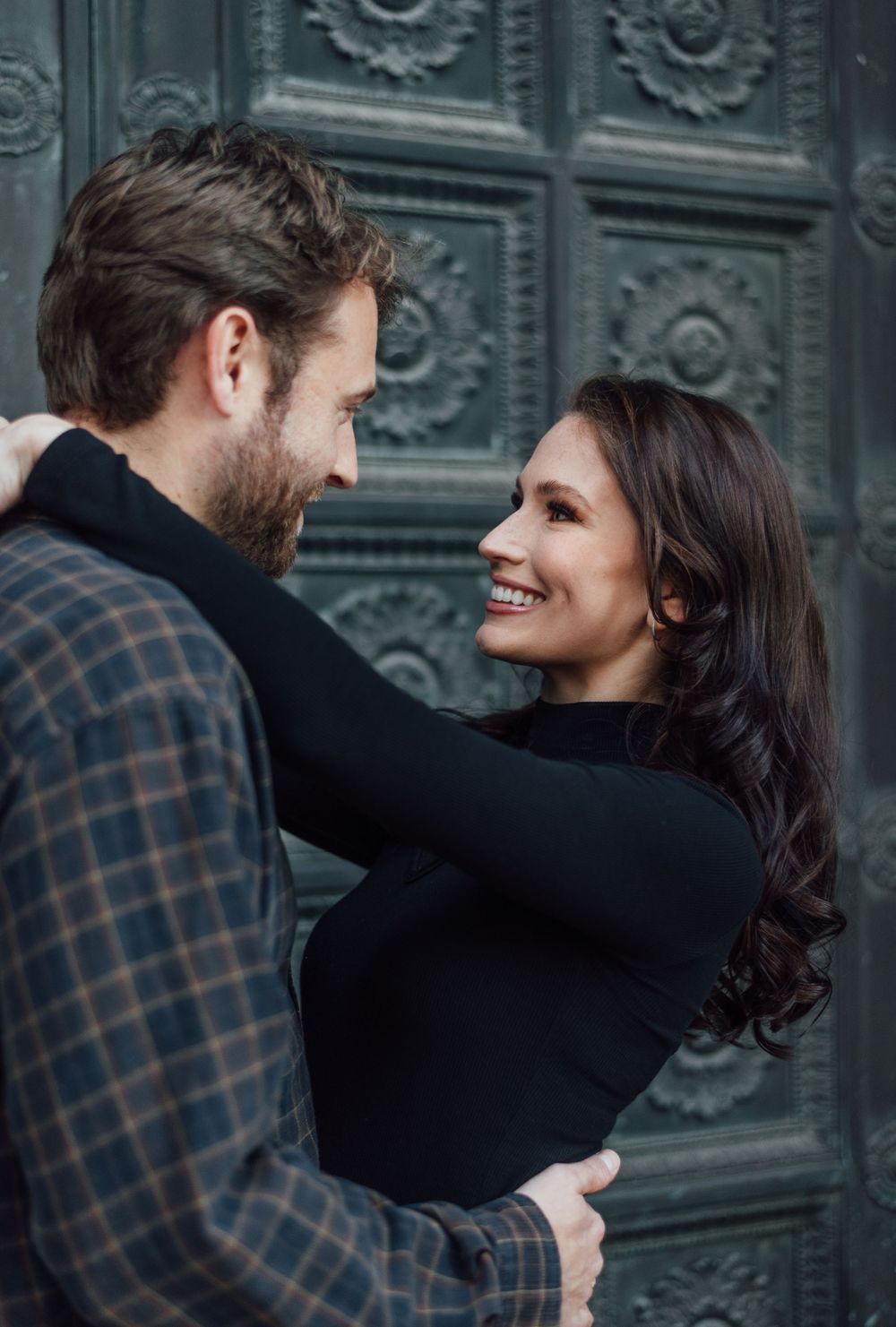 A couple embraces and smiles at each other in front of a decorative teal door with floral patterns.