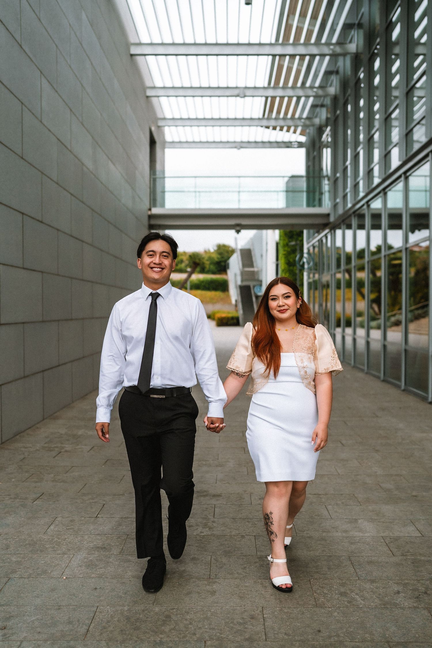 A couple walks hand in hand along a modern building corridor in formal attire.