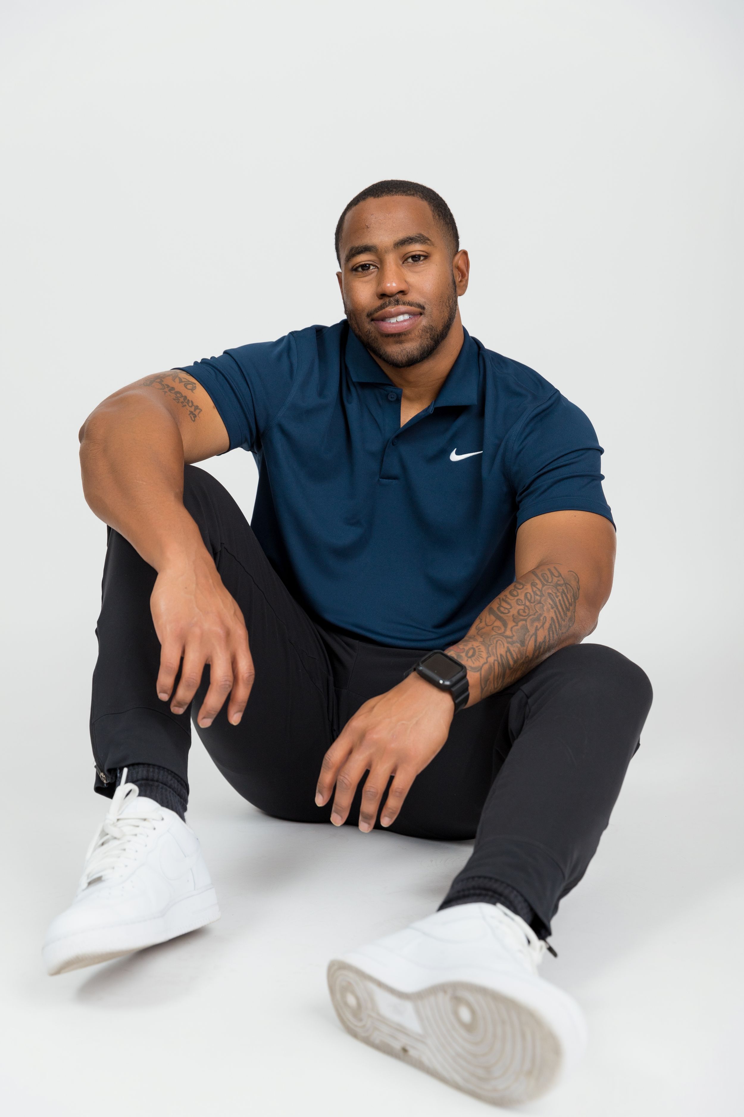 Athletic man in navy Nike polo shirt sitting casually on white studio floor.