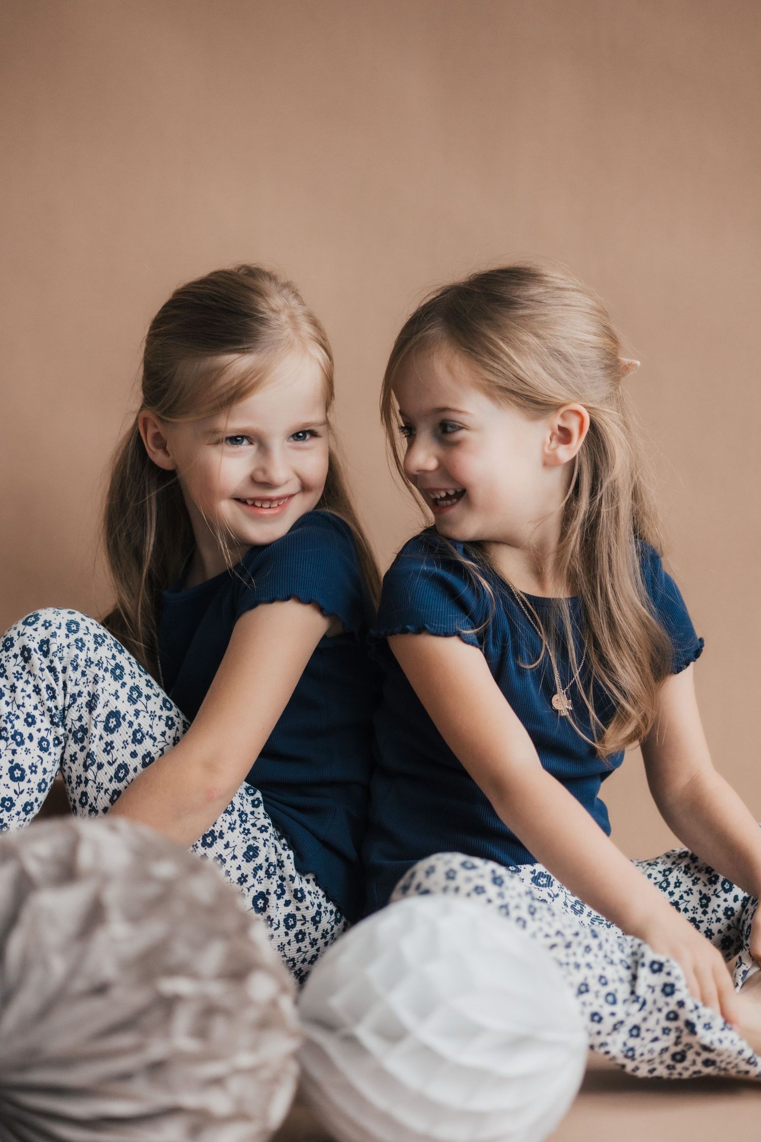 Two children in matching blue dresses share secret giggles and whispers while sitting together.