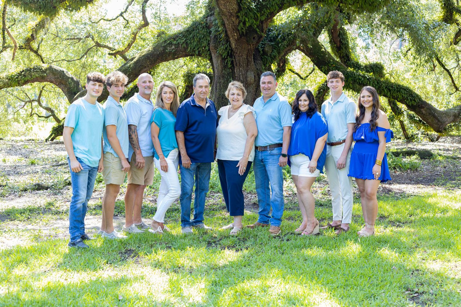 A group of people in coordinating blue and white outfits pose together under large oak trees for outdoor photos.