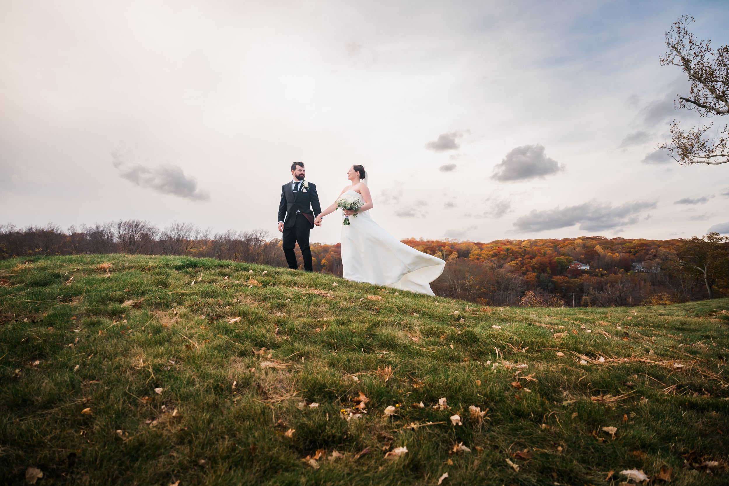 A bride and groom walking along a hilltop meadow during golden hour with dramatic clouds in the sky at Great River Golf Club.