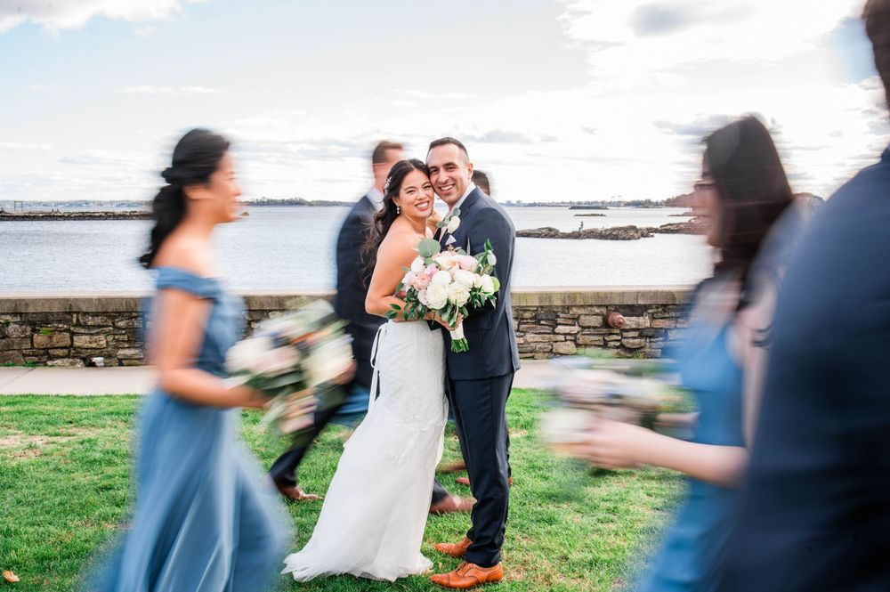 A couple shares a moment at their waterfront wedding while bridesmaids in blue dresses create motion blur around them.