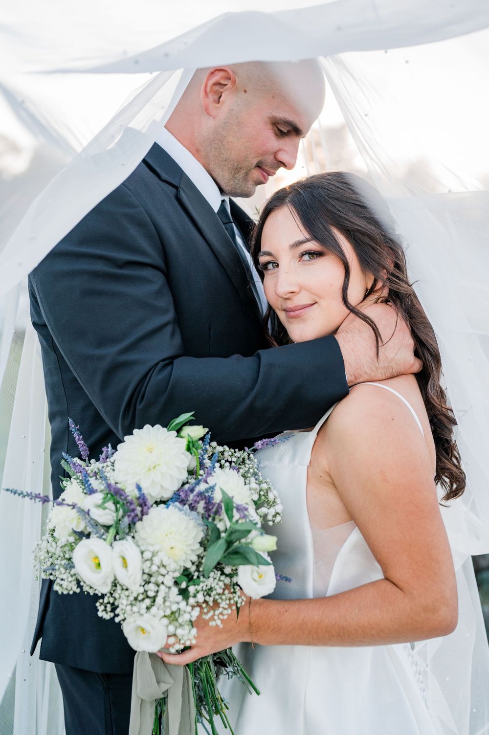Romantic wedding couple embraces under white canopy with elegant bouquet of white and purple flowers.