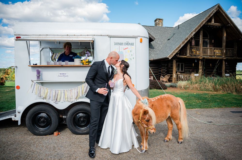 A wedding couple shares a moment beside a food truck and miniature pony at a rustic barn venue.
