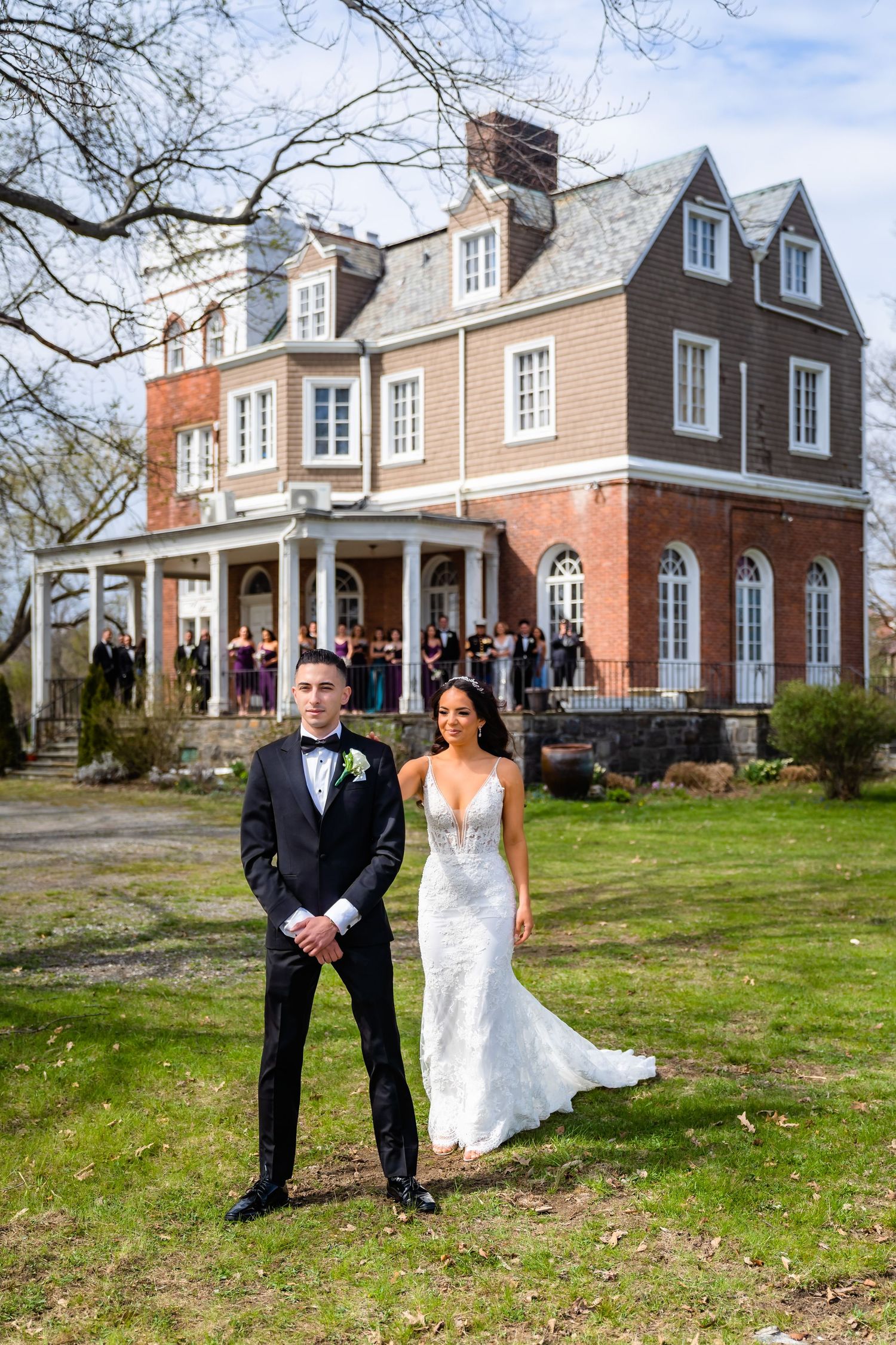 An elegant wedding couple poses in front of a historic Victorian mansion with peach and brown exterior and wraparound porch.