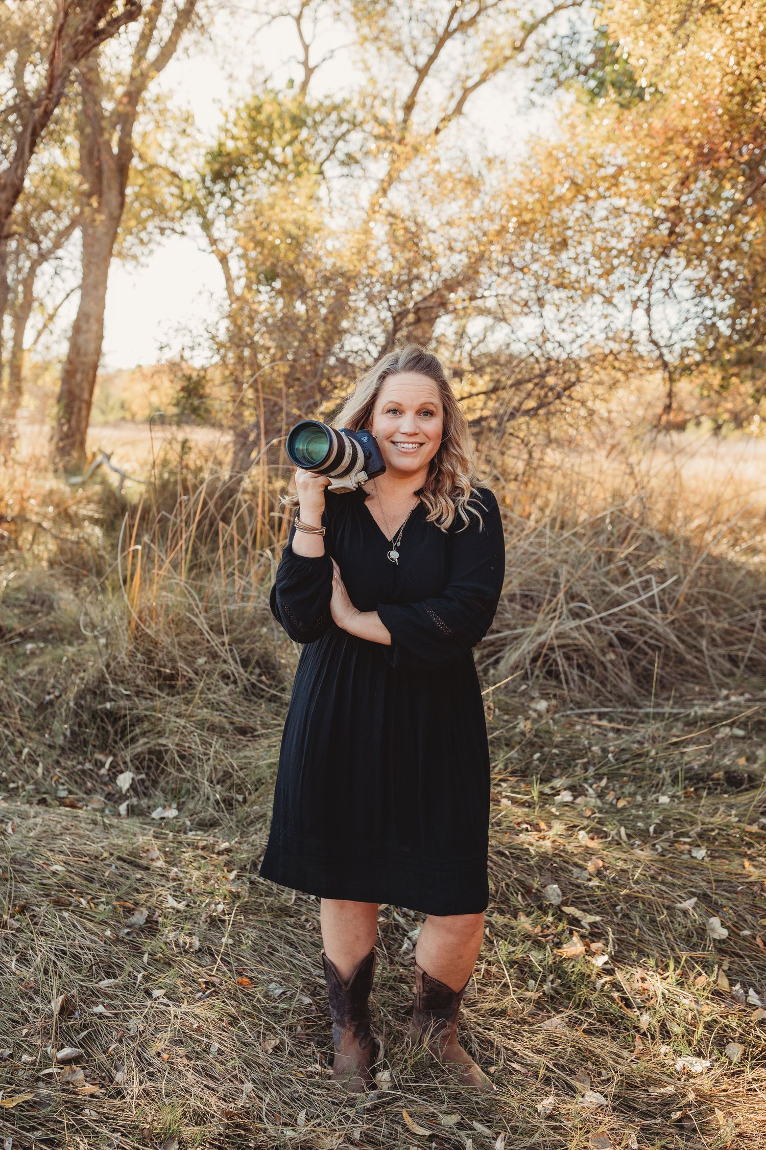 A photographer in a black dress and boots holds a camera in a sunlit autumn outdoor setting.