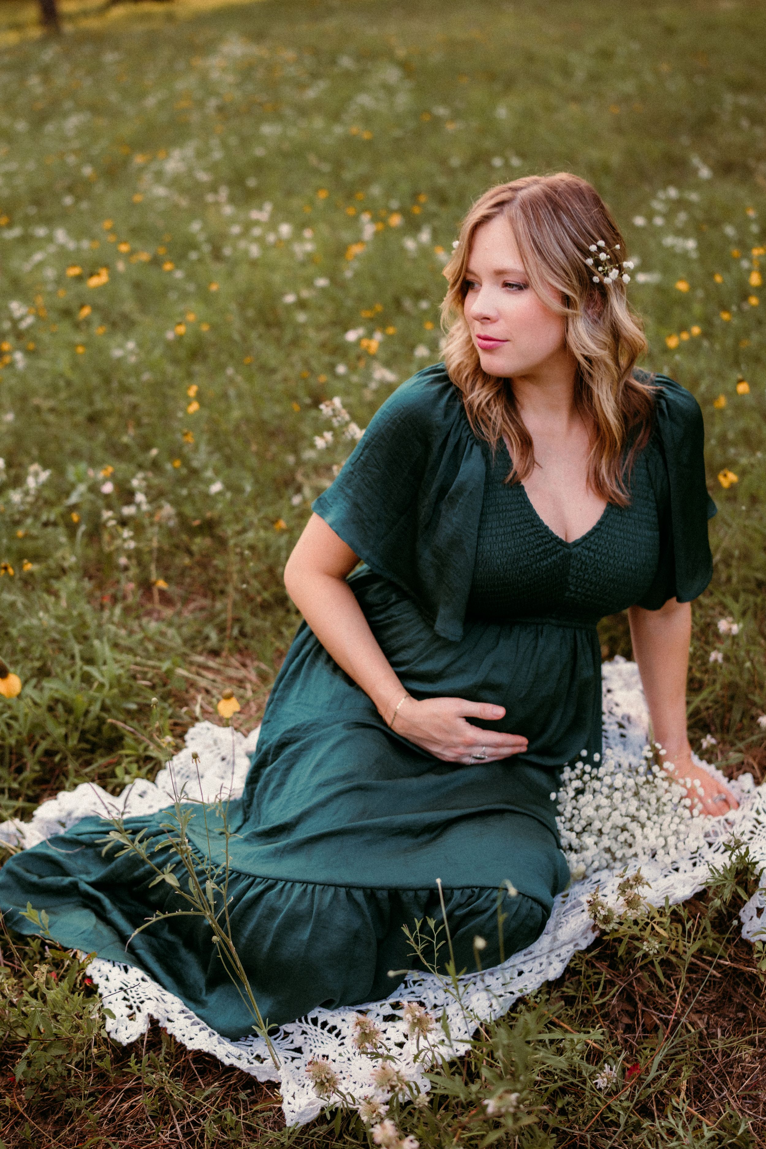 A woman in an emerald green dress sits in a wildflower meadow with delicate flowers in her wavy hair.