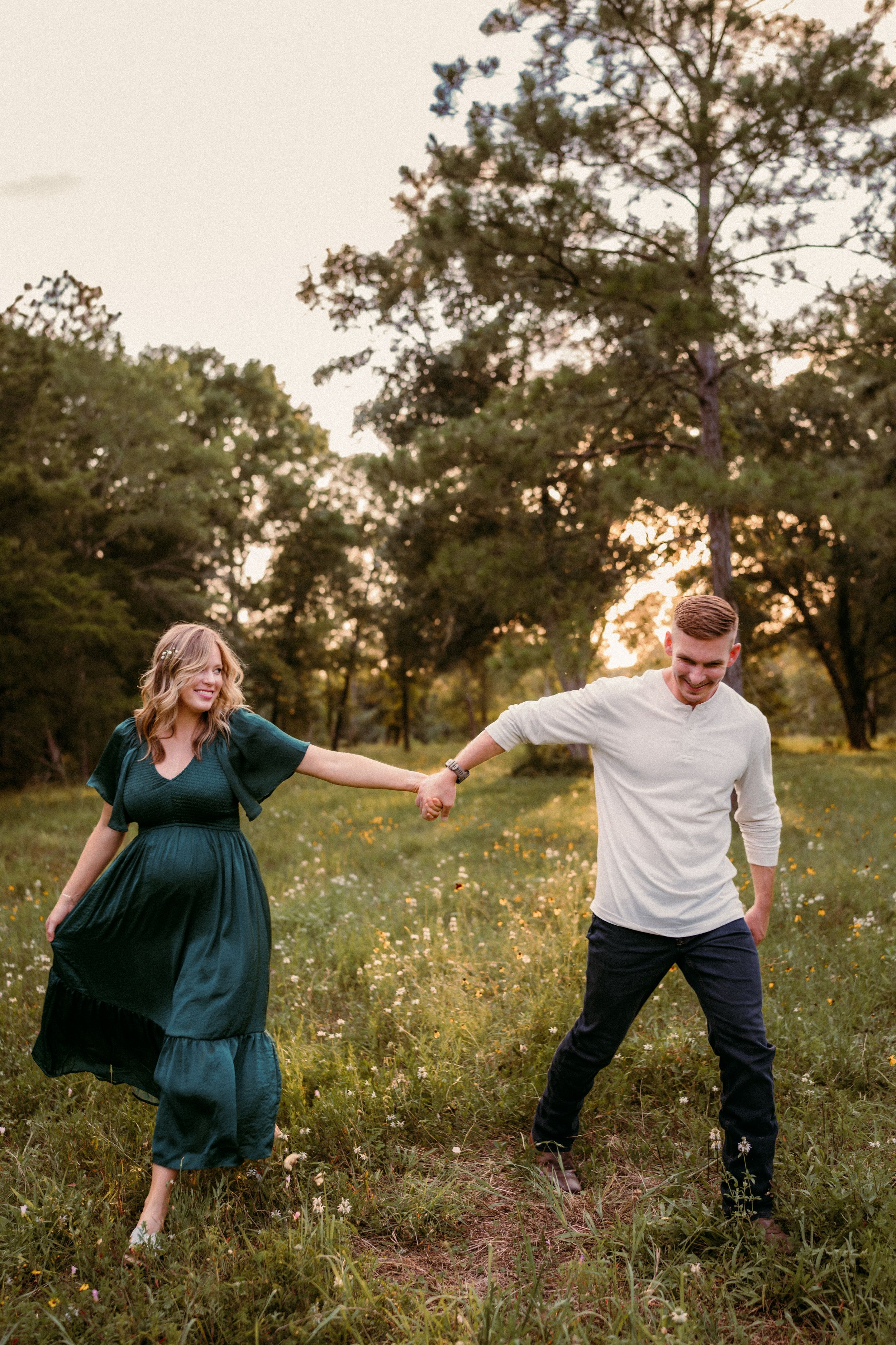 A couple holding hands in a meadow at sunset with tall pine trees in the background.