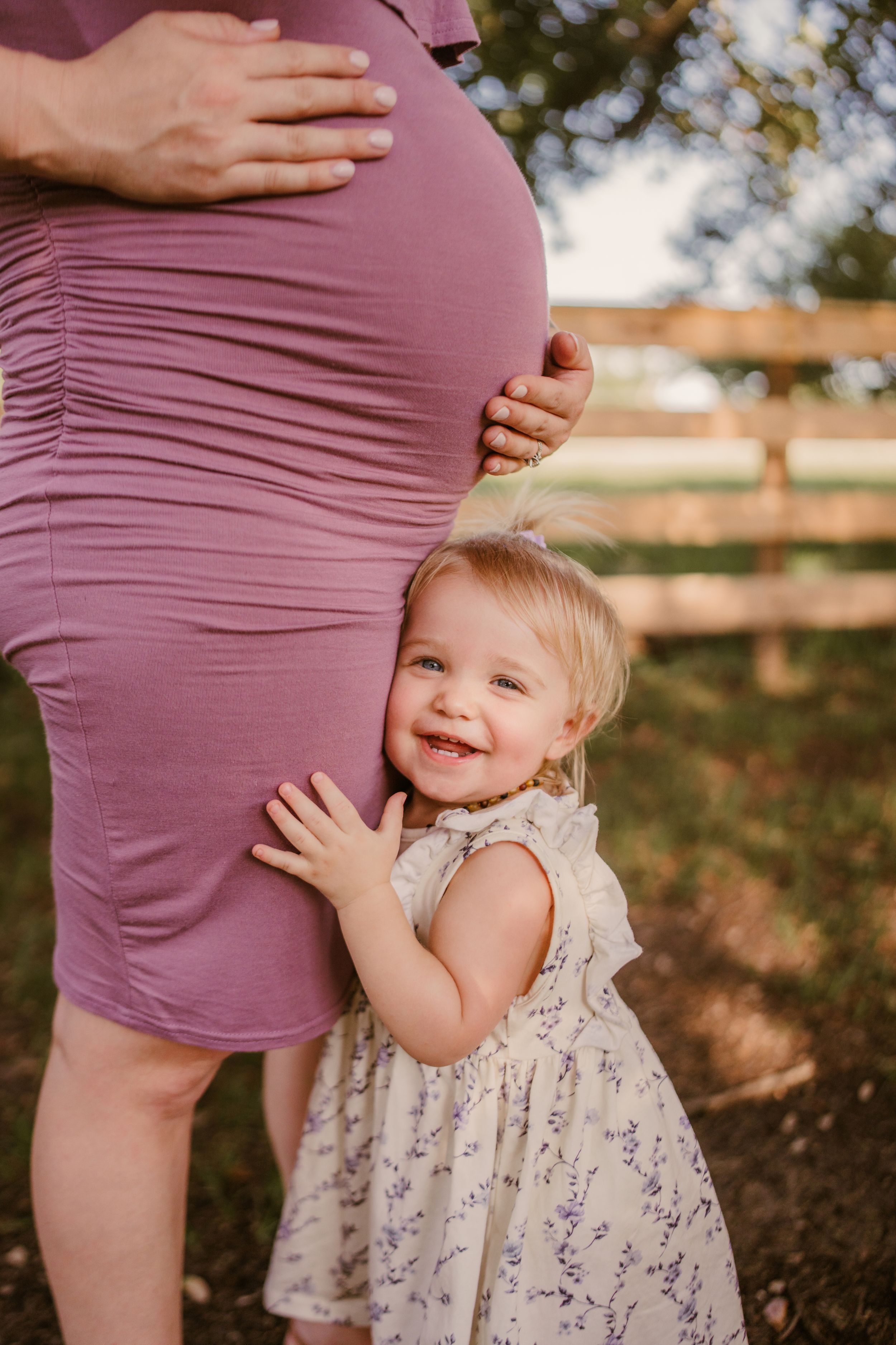 A joyful toddler in floral dress hugs a pregnant belly wearing a mauve dress outdoors near a wooden fence.