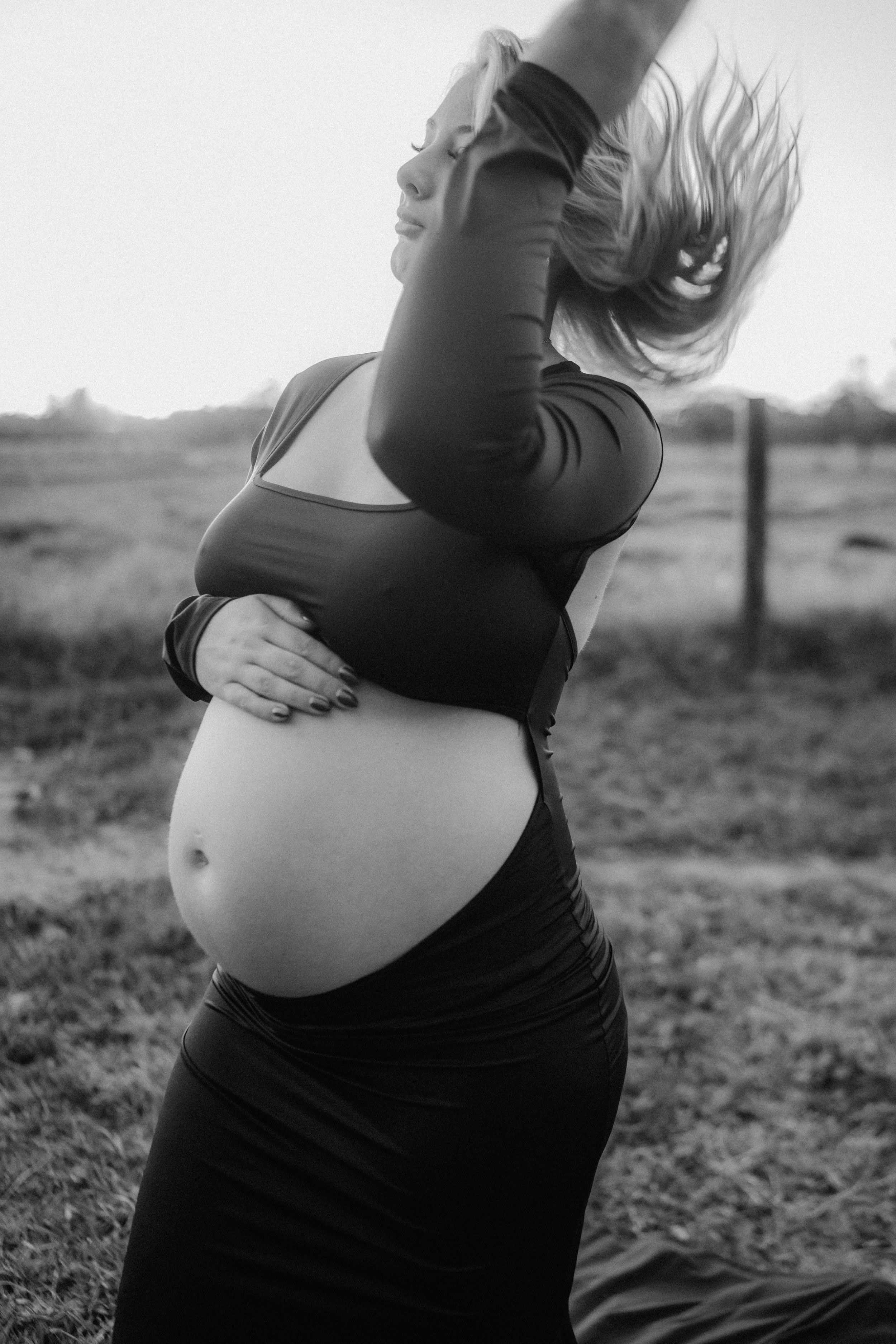 A black and white artistic maternity photo shows an expectant mother in dark clothing arching back with hair flowing in the wind.