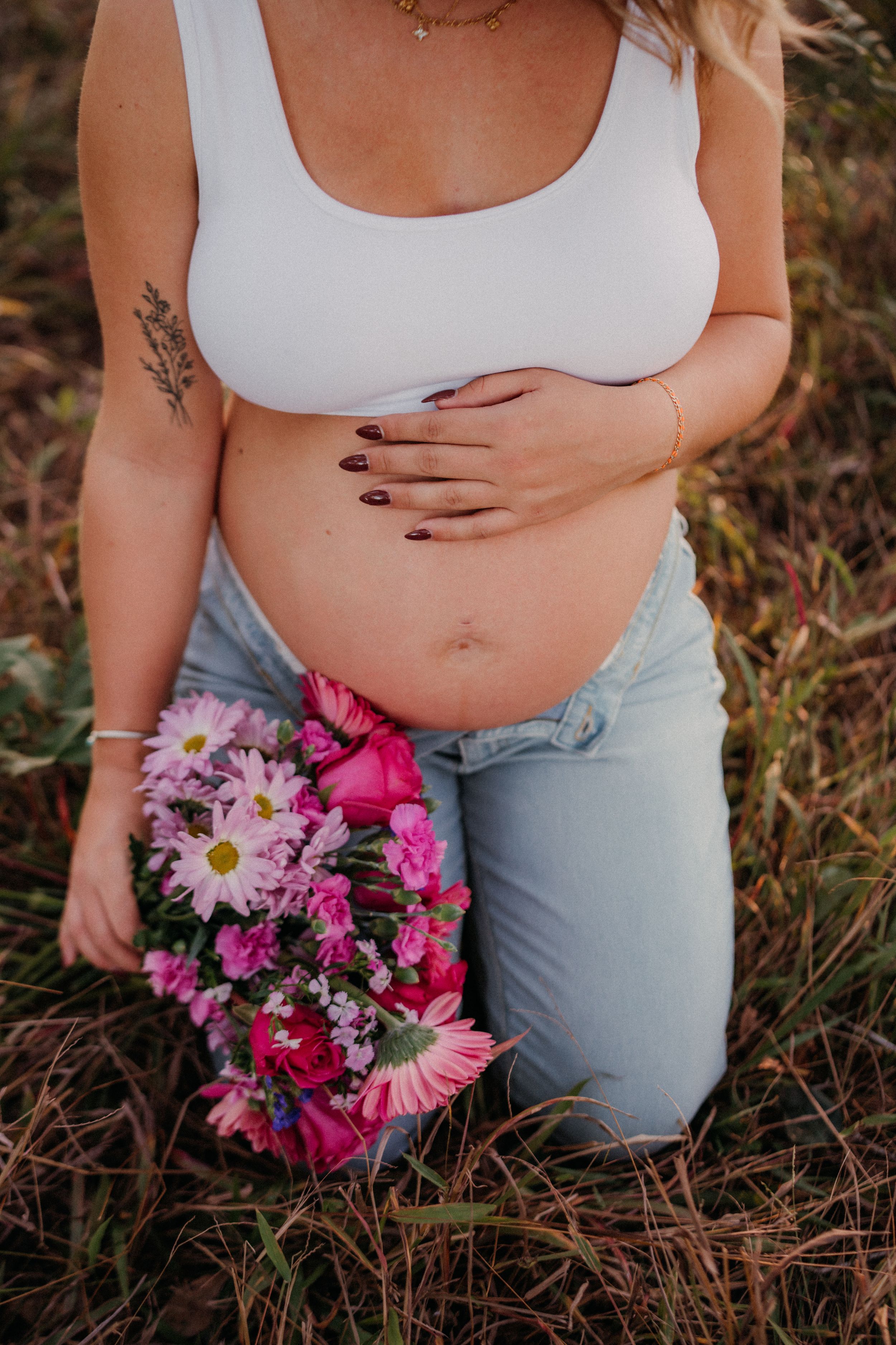 Expecting mother in white tank top and jeans holds colorful pink and purple wildflower bouquet while kneeling in grass.