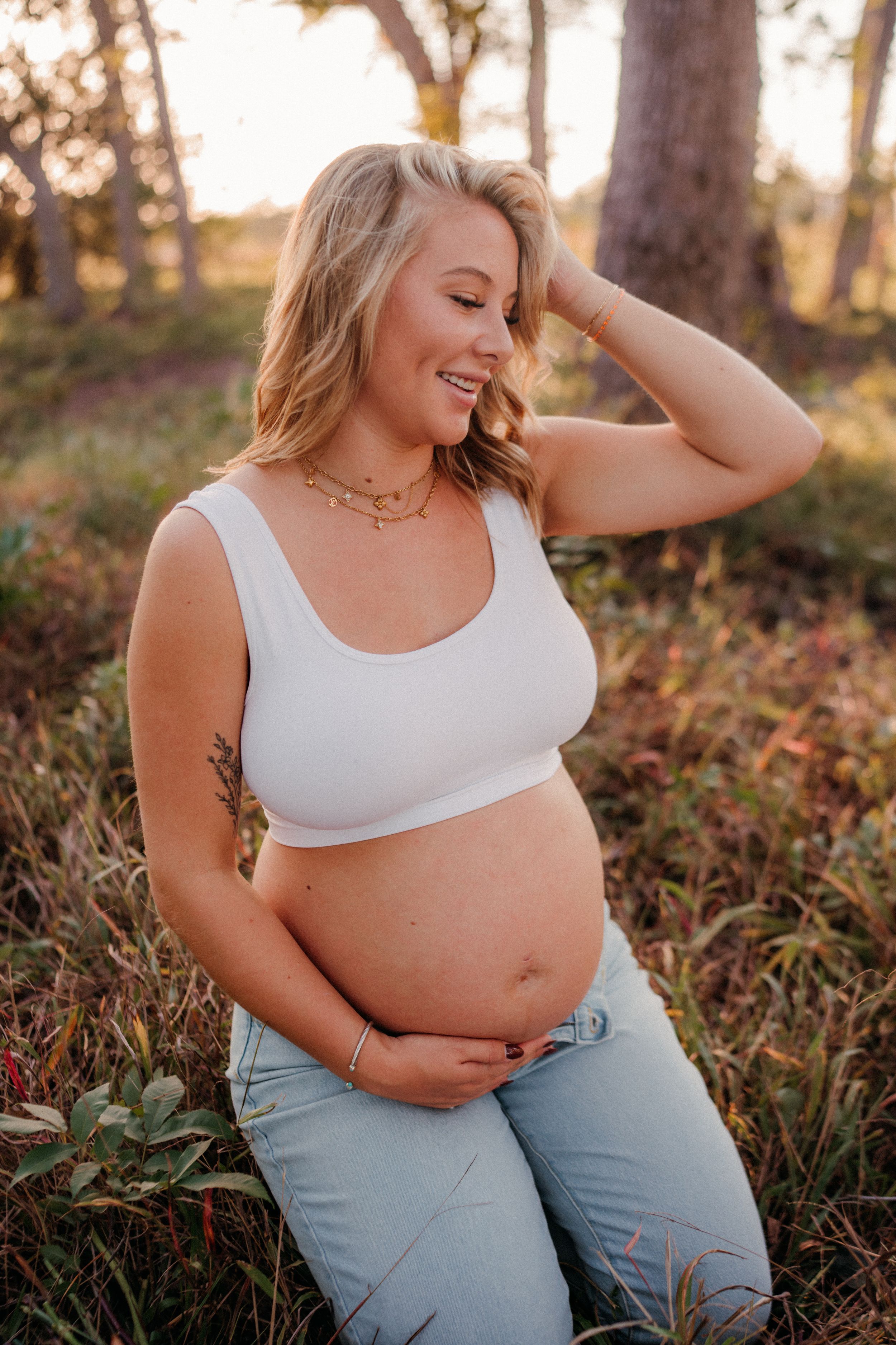 A pregnant woman wearing a white crop top and jeans poses outdoors during golden hour in a natural setting.