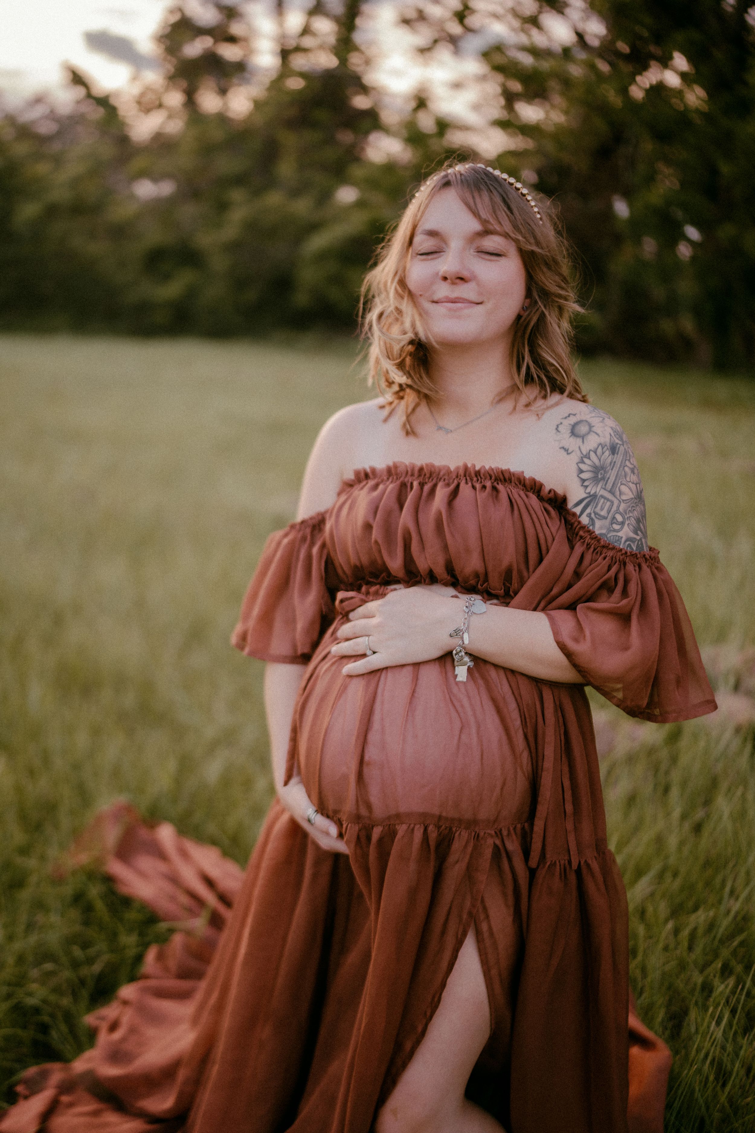 Expectant woman in flowing rust-colored dress stands peacefully in golden sunlit meadow at sunset.