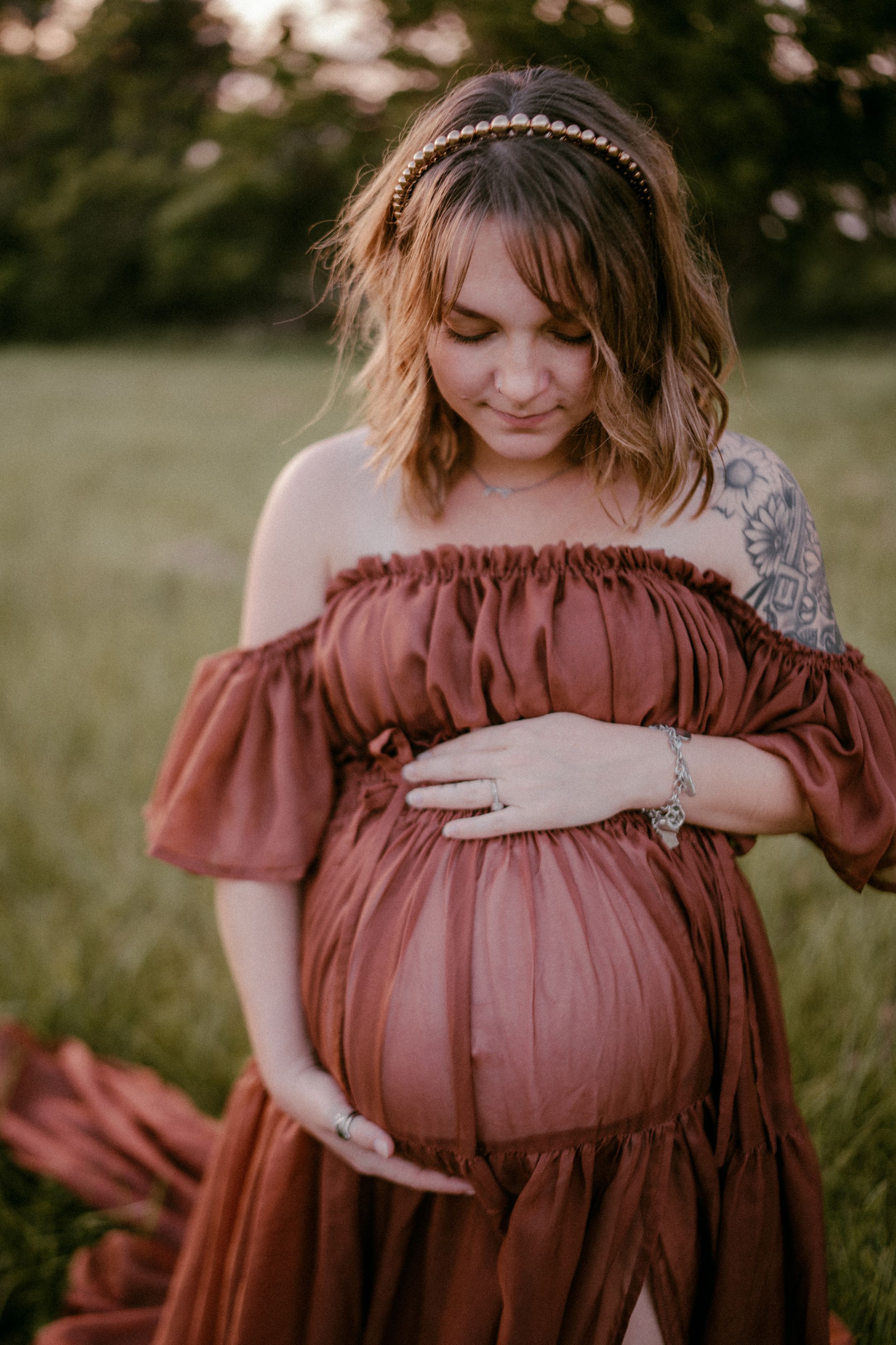 A woman in a rust-colored off-shoulder dress gently cradles her pregnant belly in a sunlit field.