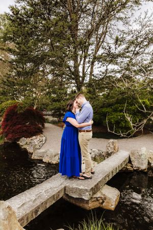 Romantic couple embracing on stone bridge over stream with lush garden backdrop.