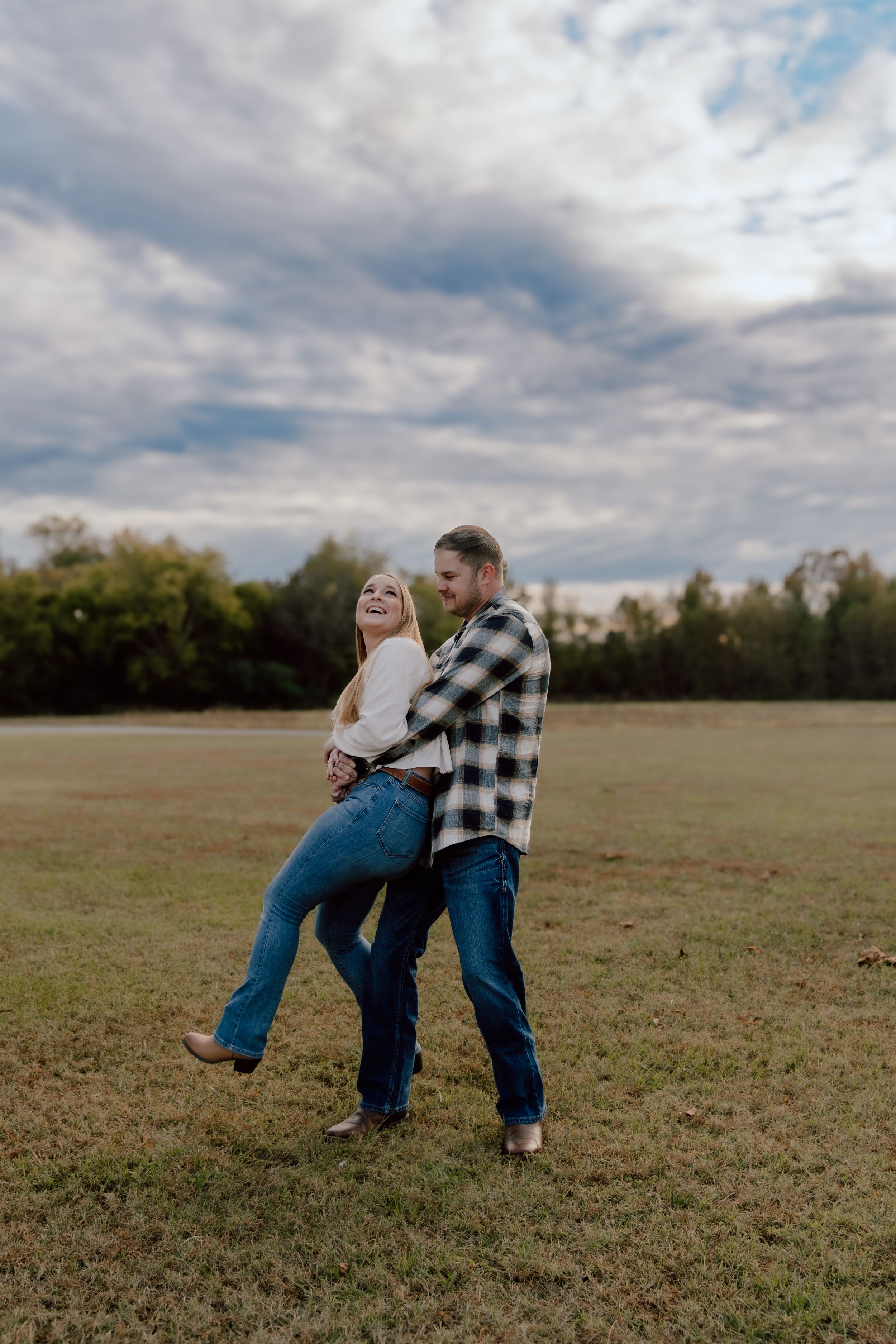 A playful couple dances and spins in a grassy field under cloudy skies, wearing casual denim and plaid.