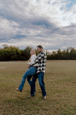 A playful couple dances and spins in a grassy field under cloudy skies, wearing casual denim and plaid.