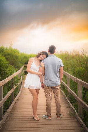 Two people standing together on a wooden boardwalk during a warm sunset with green vegetation in the background.