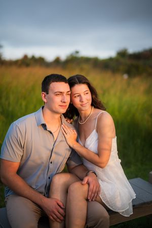 A couple sits close together on a wooden bench surrounded by tall grass during a romantic outdoor photo session at sunset.