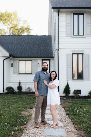 Couple smiling and embracing on a stone path in front of a white modern farmhouse with dark trim.