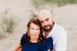 A woman in a blue dress and a bald man with a beard pose together on a sandy beach.