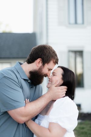 Bearded man in gray polo tenderly holds woman's face as they share a romantic moment outside a white house.