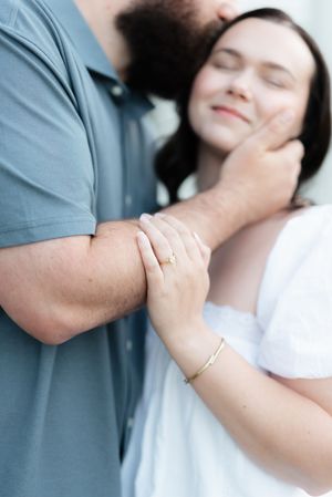 Close-up of couple embracing, woman wearing engagement ring and gold bracelet, leaning against man in gray polo.