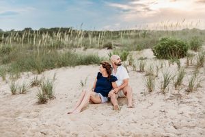 Couple sitting together on sandy beach dunes with sea oats grass at sunset.