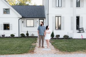 Couple standing together on a gravel path outside a white farmhouse, gazing upward on a sunny day.