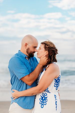 Couple embracing and almost kissing on a beach, man in blue shirt and woman in white blue floral dress, ocean behind them.