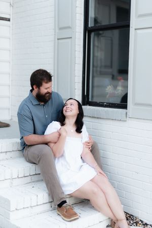 A bearded man in a blue shirt sits with a smiling woman in a white dress on white brick steps outside a building.