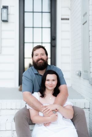 Smiling bearded man and dark-haired woman pose together on white brick porch steps of a modern home.